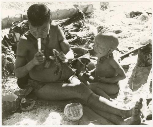 Tsekue siting with N!whakwe holding meat and a knife (print is a cropped image)