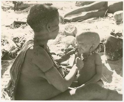 Tsekue giving N!whakwe a piece of meat, cutting it off at his lips with a knife; Oukwane in background (print is a cropped image)