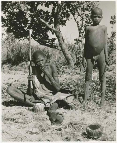 /Twikwe eating a tsama melon, !Oukabe standing beside her (print is a cropped image)