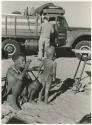 Tsekue sitting, with N!whakwe standing next to her, William Donnellan standing next to an expedition truck in the background (print is a cropped image)