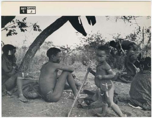 [No folder title]: Group of Ju/'hoansi sitting in a werft; left to right: /Gasa (/Qui's wife and !Naishi's daughter), /Qui (Gao's brother), //Kushay (/Gasa and /Qui's daughter) holding a stick, and other unidentified people (print is a cropped image)