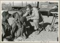 Tsekue and another person sitting, with N!whakwe standing next to them, talking with Daniel Blitz, expedition truck in the background