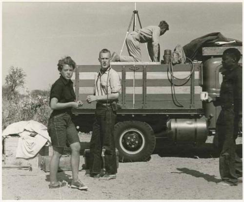 Philip walking and bringing tea to Elizabeth Marshall Thomas and John Marshall, with Daniel Blitz on his knees in an expedition truck in the background (print is a cropped image)