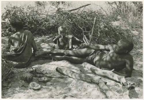 !Gai lying beside his fire, Tsekue and N!whakwe sitting beside him (print is a cropped image)