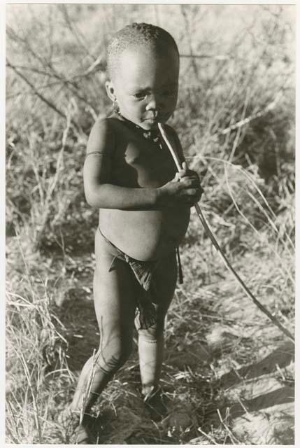 N!whakwe standing, holding a stick (print is a cropped image)