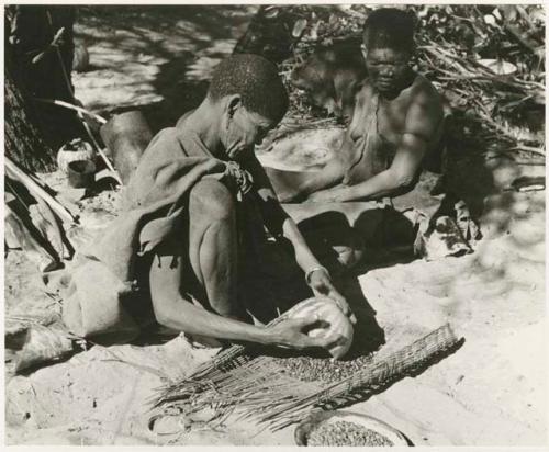 /Twikwe pouring tsama melon seeds from a rind onto her sieve, DaSi!Na sitting beside her (print is a cropped image)