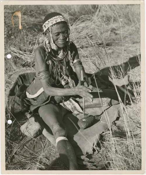 [No folder title]: "Old /Gam" sitting and drilling holes in ostrich eggshell beads with her drill (print is a cropped image)