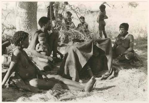 "Groups: women & children": Group of children under a kaross, Debe sitting in his father's lap, and /Naoka and Di!ai sitting (print is a cropped image)