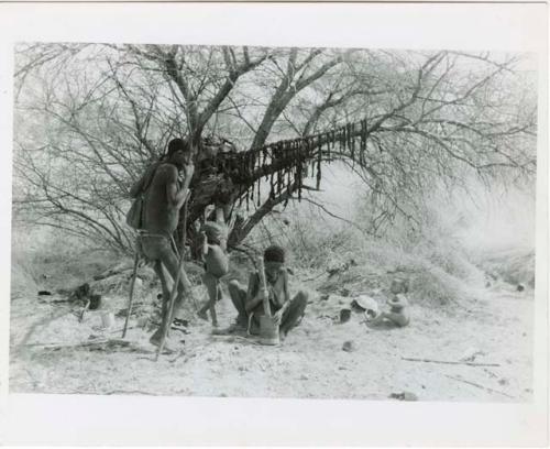 "Lame !Kham": !Kham standing with sticks as crutches, his wife pounding with a mortar and pestle, and two of his children at their dwelling place; meat he has been given hangs from a tree branch (print is a cropped image)