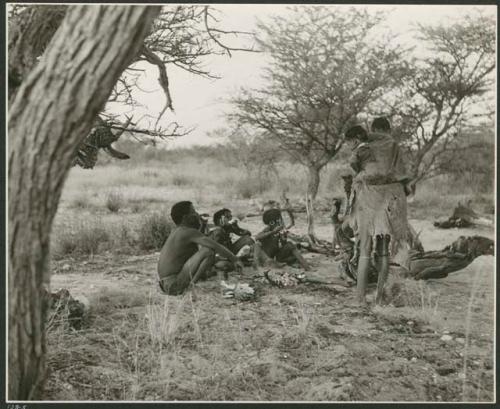 Band 1, an overnight encampment: ≠Toma with a group of people standing and sitting, one is eating a tsama melon (print is a cropped image)