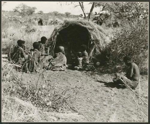 "Band 2, the group visiting /Qui Hunter": Group of people sitting in front of a skerm, including "Old /Gasa," //Khuga (wife of /Naishi), "Little /Qui" and "Old Gau" from Band 1 (print is a cropped image)