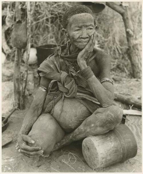 "Band 1, Old N/aoka 413 Mo. of !U, etc.": Old N/aoka, mother of !U, sitting by a fire, her knee over a mortar (print is a cropped image)