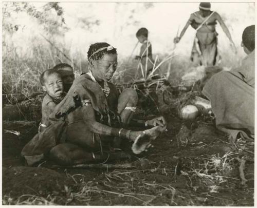 [No folder title]: Di!ai preparing wood for a fire, and her child sitting next to her crying (print is a cropped image)