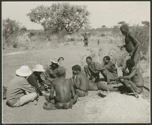 Family in Nai Nai: Laurence Marshall, Lorna Marshall, and Kernel Ledimo, wearing a hat with a feather, speaking with several men sitting on the ground including ≠Toma and ≠Gao (Khwo//o-/Gasa's husband); "Old Demi" standing to the right (print is a cropped image)