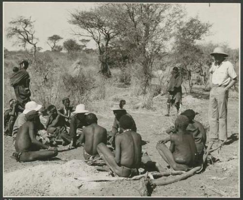 Family in Nai Nai: Laurence Marshall, Lorna Marshall, and Kernel Ledimo wearing a hat with a feather speaking with several men sitting on the ground including ≠Toma, ≠Gao (Khwo//o-/Gasa's husband), and "Old Demi"; Hans Ernst standing to the right and unidentified women in the background (print is a cropped image)