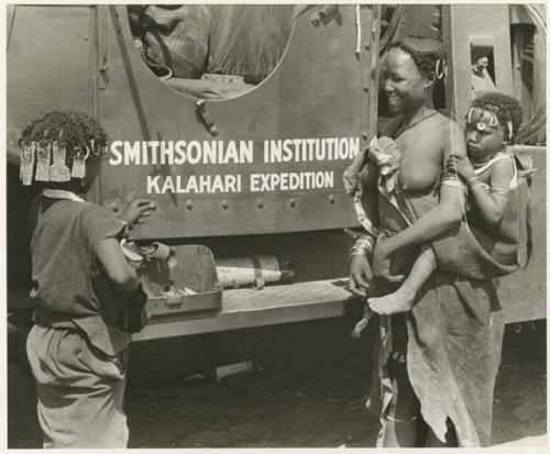 Family in Nai Nai: N!ai and a woman standing in front of one of the expedition's GMC trucks with "Smithsonian Institution Kalahari Expedition" showing (print is a cropped image)