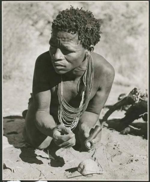 [No folder title]: ≠Toma crouching and holding a soapstone pipe (print is a cropped image)