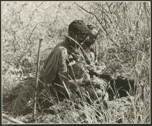 [No folder title]: Woman sitting in the grass by a hole she made digging up a root (print is a cropped image)