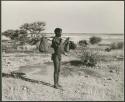 [No folder title]: /Ti!kay carrying bags on a carrying stick and an assegai, with the pan in the background (print is a cropped image)