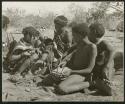 [No folder title]: Group of people sitting, including N!ai (left), //Khuga (middle), and /Gishay playing the //guashi (right) (print is a cropped image)