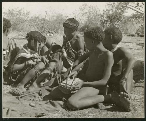 [No folder title]: Group of people sitting, including N!ai (left), //Khuga (middle), and /Gishay playing the //guashi (right) (print is a cropped image)