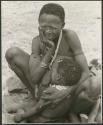 [No folder title]: Khuan//a (Gau's second wife) sitting on the ground with her chin in her hand and holding an unidentified child against her (print is a cropped image)