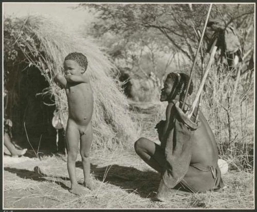 [No folder title]: /Ti!kay sitting, with his hunting equipment next to him, and !Nani standing beside him (print is a cropped image)