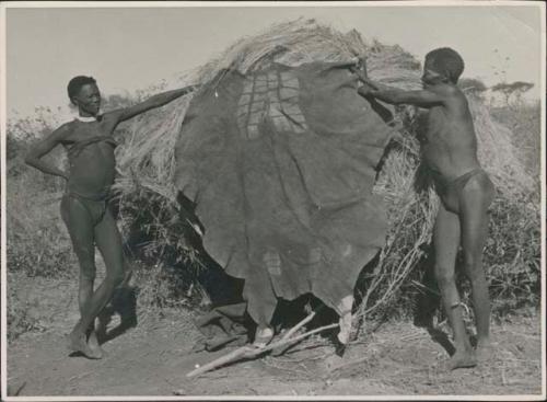 "Old ≠Toma" of Band 2 and Gau holding a kaross up against a skerm, showing the pattern on it created by scraping part of the hair off the animal skin