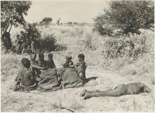 Group of women sitting in the dance circle and a man lying in a trance, with a group of men sitting in the shade in the background
