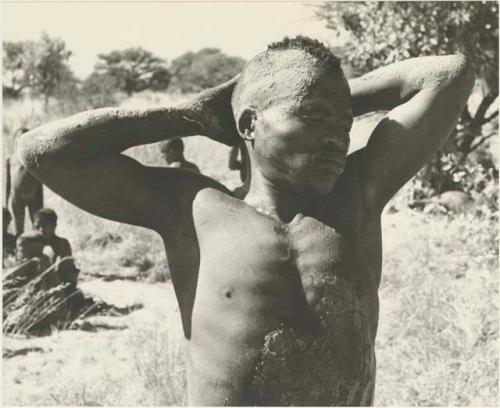 Medicine man putting his arms behind his head, half figure standing
