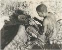 !Gai seated, cutting another man's hair, seen from behind

