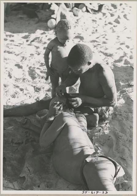 !Gai seated, cutting another man's hair, with a child standing next to him


