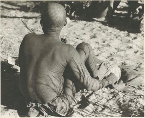 Oukwane seated and playing a musical bow, seen from behind

