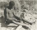 !Gai shaping an arrow point made of bone, sitting with his legs over a log




