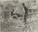 One boy playing a sanza and another boy dancing in a circle with dance rattles tied to his ankles


