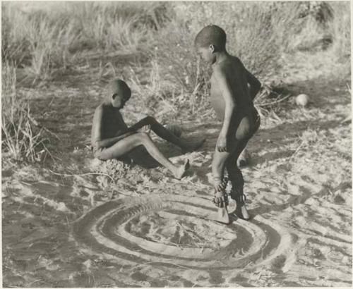 One boy playing a sanza and another boy dancing in a circle with dance rattles tied to his ankles


