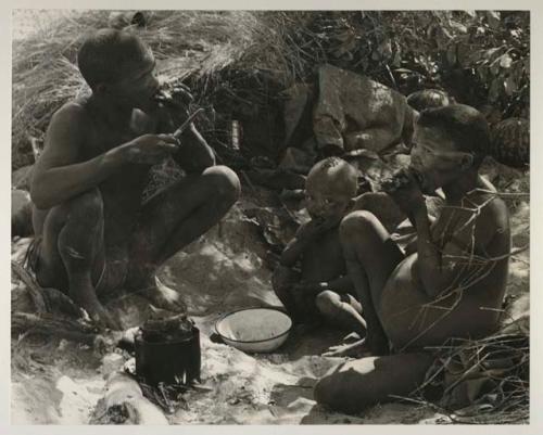 !Gai, Tsekue, and N!whakwe sitting by their fire and eating meat, with meat cooking in a tin on it

