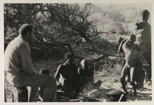 Oukwane sitting by his fire, a boy carrying N!whakwe nearby, and William Donnellan sitting and watching them
