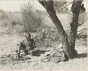 Oukwane seated, playing a bow under a tree, distant view

