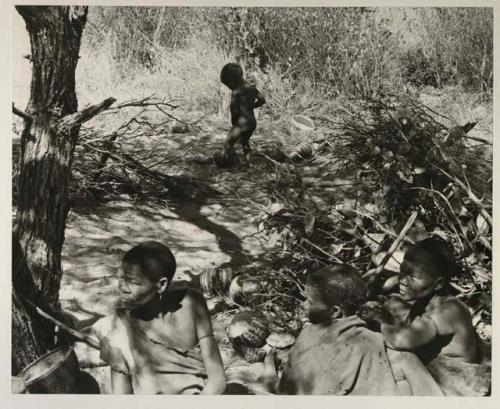 Group of women sitting, and N!whakwe urinating in a tsama melon rind in the background
