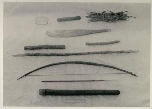 /Twikwe's possessions, including (top to bottom, left to right) a piece of wood hollowed in center used to trip iron traps, a small stick, a bundle of sharpened sticks used to peg down a hide to stretch it, a fire paddle, 2 sticks, a digging stick, a bow, arrow with a bone point shaped like a needle, and a quiver, with a ruler for measurement