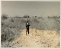 N!whakwe standing in the grass drinking from a tin
