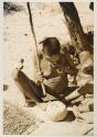 /Twikwe eating a piece of tsama melon, with a pile of seeds on a mat on the ground near her
