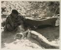 Tsekue nursing N!whakwe beside a fire, with !Gai lying beside them under his big kaross


