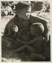 Tsekue nursing N!whakwe, seated beside a fire
