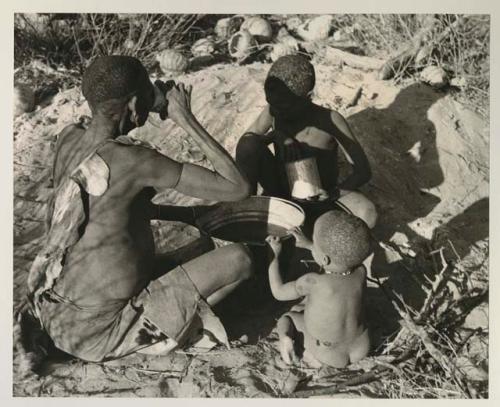 !Twikwe, N!whakwe, and a little child sitting around a bucket of water; Twikwe is drinking from a tin


