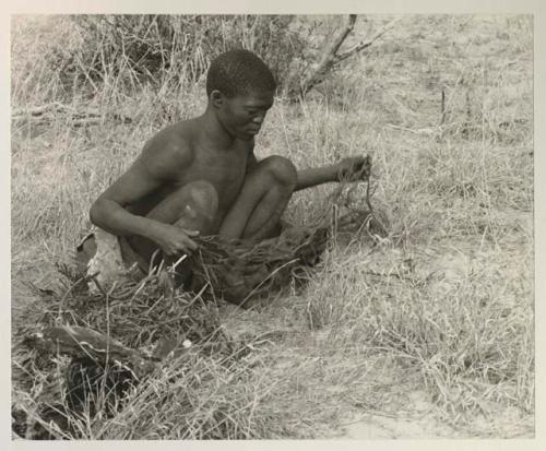 !Gai squatting in the grass holding a carrying net
