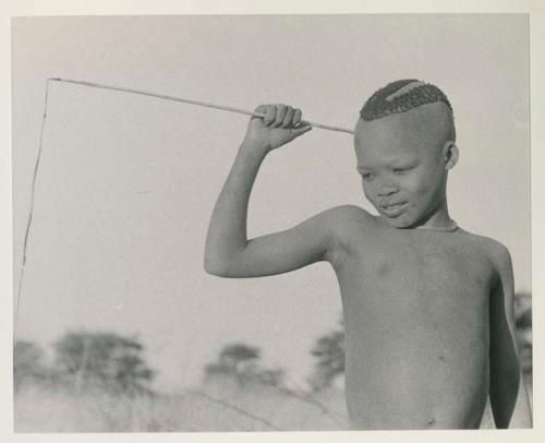 Boy holding a stick, to which a feather is attached by a thong, to make a noise

