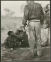 [No folder title]: Woman nursing a baby and having ointment put in her eyes by Lorna Marshall, with Laurence Marshall and other expedition members watching (print is a cropped image)