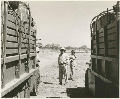 [No folder title]: Laurence Marshall and Lorna Marshall standing between two expedition trucks (print is a cropped image)